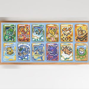 Puede incluir: Una obra de arte en punto de cruz enmarcada con diez diseños de cartas Pokémon coloridas. Cada carta muestra un personaje diferente, con detalles vibrantes y una estética pixelada. El marco es marrón claro y el fondo es azul claro.