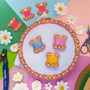Puede incluir: Un bastidor de punto de cruz con un borde floral rosa, que contiene carretes de hilo azul, rosa y amarillo. Las margaritas decorativas y los carretes de hilo están dispersos alrededor del bastidor. También se ven unas tijeras y unos alicates.