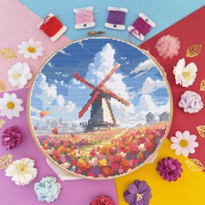 Puede incluir: Un aro de bordado de punto de cruz colorido que representa un molino de viento en un campo de flores. El molino de viento es rojo y blanco con un cielo azul y nubes blancas en el fondo.