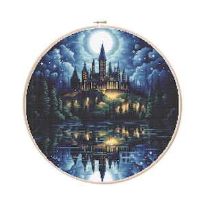 May include: A cross-stitch embroidery of a castle scene at night, set in a wooden hoop. The castle is illuminated with warm lights, reflected in the water below. The sky is dark blue with a full moon and stars.