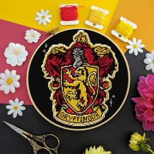 Puede incluir: Un aro de bordado negro y dorado con un diseño de punto de cruz del escudo de la casa Gryffindor de la serie Harry Potter. El escudo presenta un león, una espada y las palabras "Gryffindor" en una pancarta.