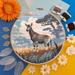 May include: A cross-stitch embroidery pattern of an okapi, a mammal native to the Democratic Republic of Congo, standing in a grassy field with a blue sky and a tree in the background.