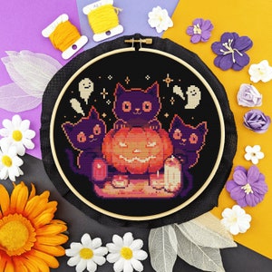 Pode incluir: Um aro de bordado preto com fundo preto e um desenho de ponto cruz colorido de três gatos pretos e uma abóbora de Halloween. Os gatos estão sentados em volta da abóbora de Halloween, que é laranja e tem uma cara assustadora.