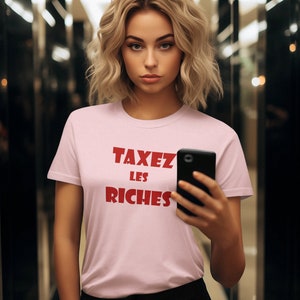 May include: A woman wearing a pink t-shirt with the text "TAXEZ LES RICHES" in red letters. She is holding a black smartphone in her right hand.