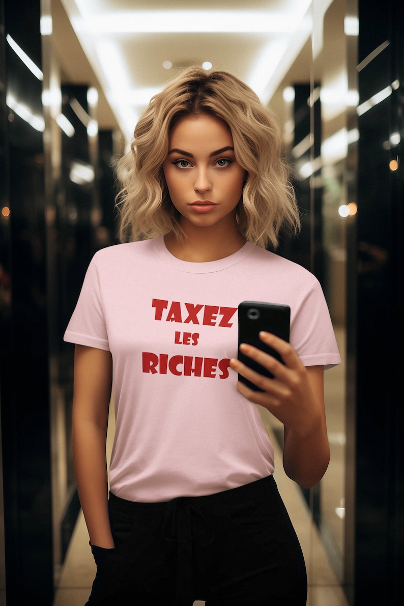May include: A woman wearing a pink t-shirt with the text "TAXEZ LES RICHES" in red letters. She is holding a black smartphone in her right hand.