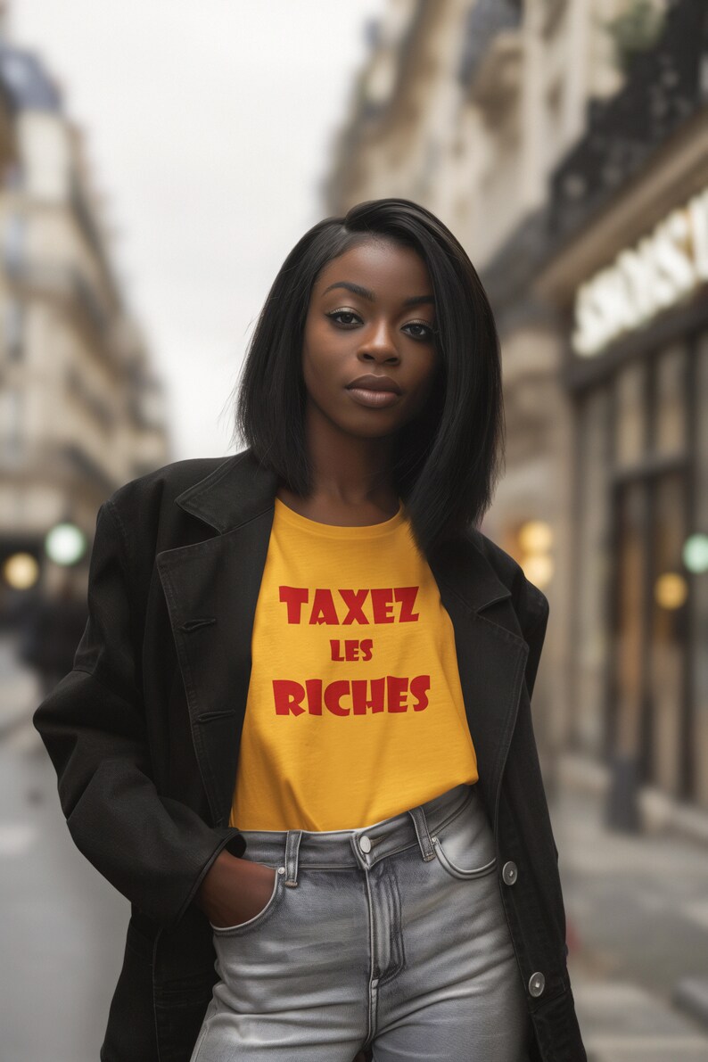 May include: A woman wearing a black jacket over a yellow t-shirt with the text "TAXEZ LES RICHES" in red. She is standing in a city street.