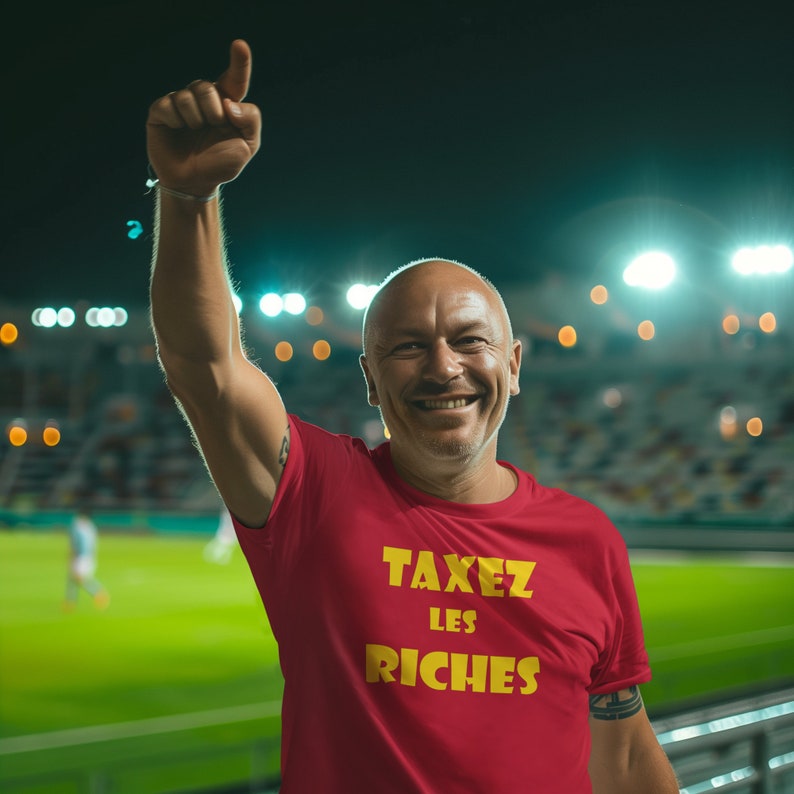 May include: A man wearing a red t-shirt with the text "TAXEZ LES RICHES" in yellow letters. He is smiling and raising his right hand in the air.