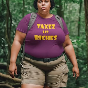 May include: A woman wearing a purple t-shirt with the text "TAXEZ LES RICHES" in yellow letters. She is wearing khaki shorts and a black belt. She is standing in a forest.