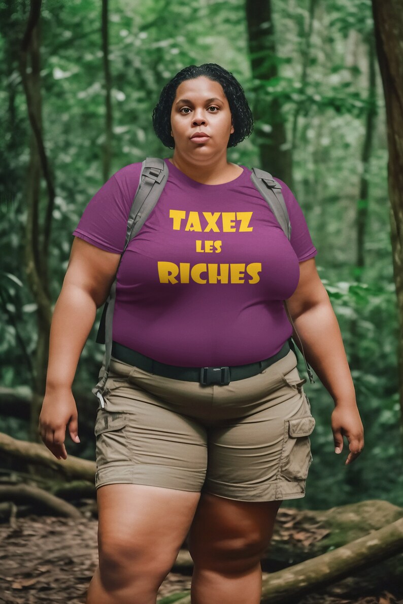 May include: A woman wearing a purple t-shirt with the text "TAXEZ LES RICHES" in yellow letters. She is wearing khaki shorts and a black belt. She is standing in a forest.