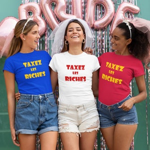 May include: Three friends wearing matching t-shirts with the text "Taxez Les Riches" in yellow and red letters. The shirts are blue, white, and red. The friends are smiling and posing for a photo.