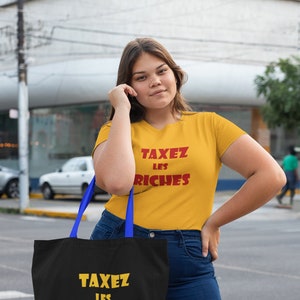May include: A person wearing a yellow t-shirt with the text "TAXEZ LES RICHES" in red. They are holding a black tote bag with the same text in yellow.