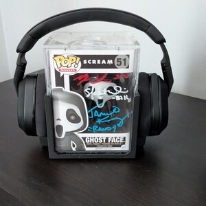 May include: A Funko Pop Movies Ghost Face vinyl figure, number 51, encased in a clear protective display box with multiple signatures in red, black, and blue ink, positioned behind a pair of black over-ear headphones on a dark surface.