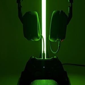 Op de afbeelding: Zwarte gaming headset stand met een ingebouwd controller oplaadstation. De standaard heeft een verticaal groen licht en wordt verlicht met een groene gloed. De headset hangt aan de bovenkant en een controller rust aan de basis.