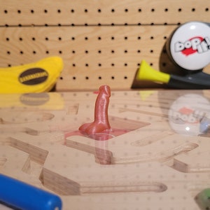 May include: A wooden maze game with a pink plastic piece in the center. The maze is made of light wood and has a light brown finish. There is a yellow banana and a blue toy in the background.