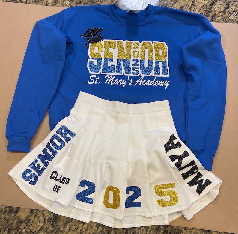 May include: A white pleated skirt with blue and gold glitter lettering that says "Senior Class of 2025" and "Mary's" on the sides. A blue sweatshirt with the text "Senior 2025" and "St. Mary's Academy" is also pictured.