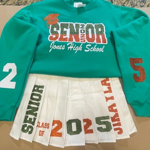May include: A white pleated skirt with green, orange, and white glitter lettering that says "Senior Class of 2025" and "Jikai La". A green sweatshirt with the text "Senior 2025 Jones High School" and the numbers "2" and "5" on the sleeves.