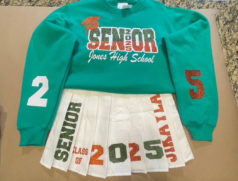 May include: A white pleated skirt with green, orange, and white glitter lettering that says "Senior Class of 2025" and "Jikai La". A green sweatshirt with the text "Senior 2025 Jones High School" and the numbers "2" and "5" on the sleeves.