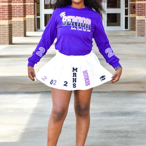 May include: A young woman wearing a purple long-sleeved shirt with the text "Senior Martin Road High School" and a white pleated skirt with the text "MRHS Senior 202. She is wearing a blue graduation cap and white heels.