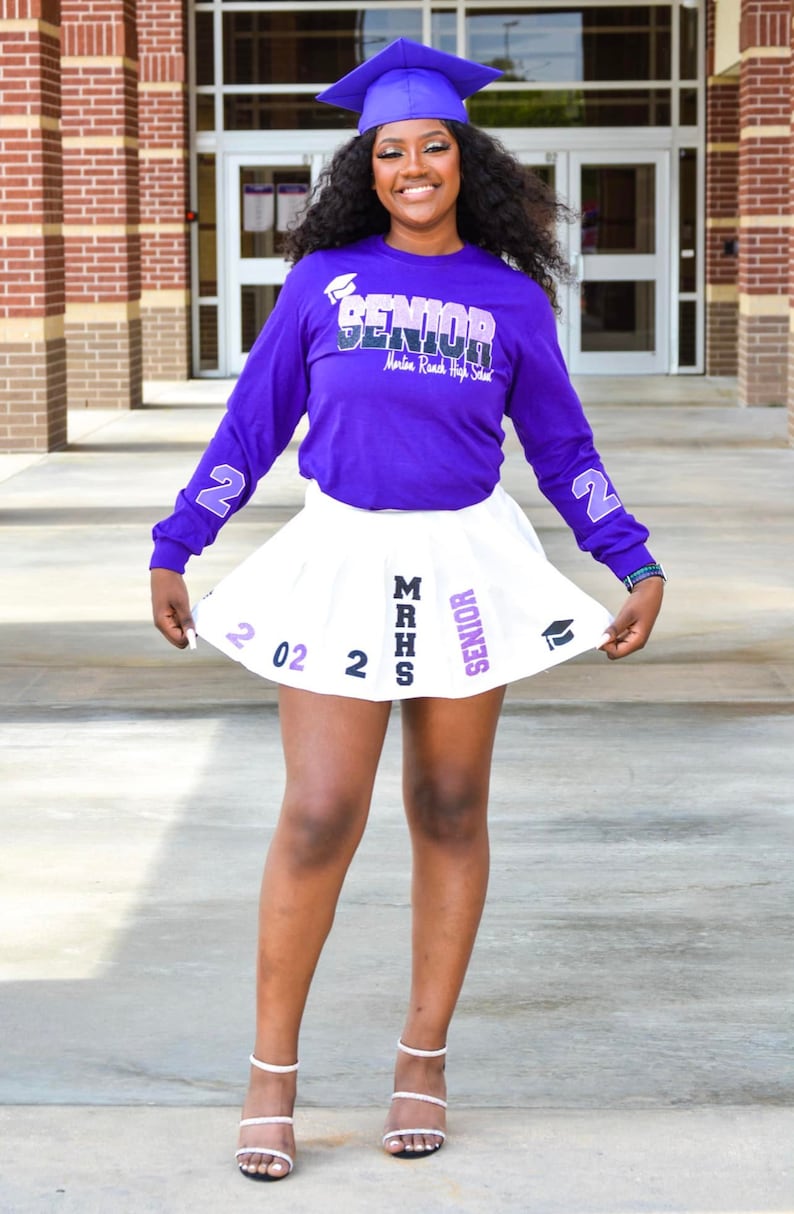 May include: A young woman wearing a purple long-sleeved shirt with the text "Senior Martin Road High School" and a white pleated skirt with the text "MRHS Senior 202. She is wearing a blue graduation cap and white heels.