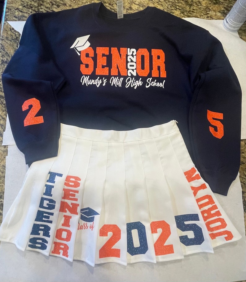 May include: A white pleated skirt with the words "TIGERS", "SENIOR", "Class of 2025", and "JORDYN" printed on it in orange and blue glitter. The skirt is paired with a navy blue sweatshirt with the words "SENIOR 2025" and "Mundy's Mill High School" printed on it in orange glitter.