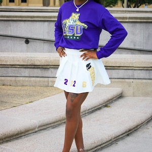 May include: A woman wearing a purple long-sleeved shirt with a tiger graphic and the text "LSU TIGERS". She is also wearing a white pleated skirt with the text "TIGERS" and "2026" on it. She is standing on a set of stairs.