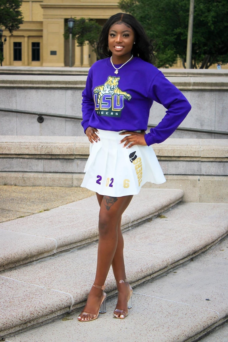 May include: A woman wearing a purple long-sleeved shirt with a tiger graphic and the text "LSU TIGERS". She is also wearing a white pleated skirt with the text "TIGERS" and "2026" on it. She is standing on a set of stairs.