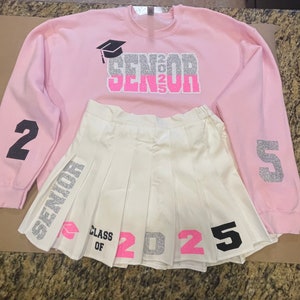 May include: Pink sweatshirt with silver glitter text that says "Senior 2025" and a graduation cap graphic. The sweatshirt has black numbers 2 and 5 on the sleeves. A white pleated skirt with silver glitter text that says "Senior" and "Class of 2025" with pink numbers.