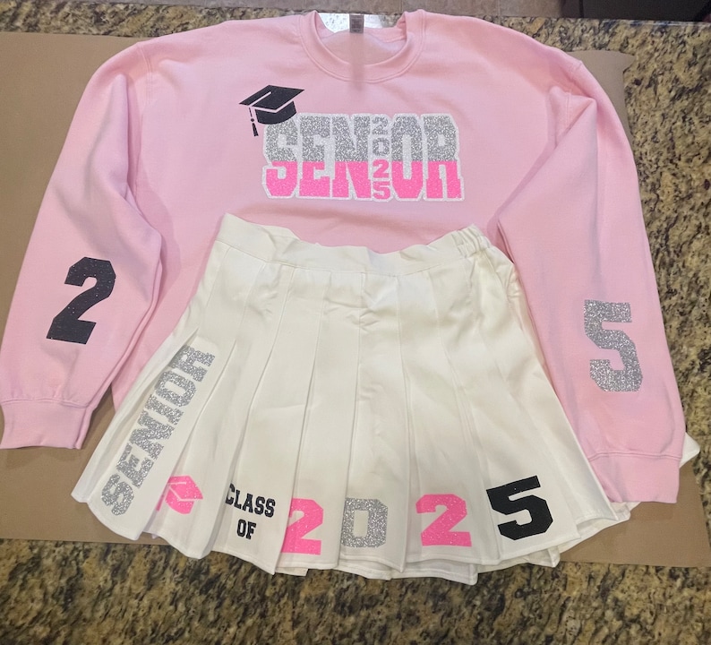 May include: Pink sweatshirt with silver glitter text that says "Senior 2025" and a graduation cap graphic. The sweatshirt has black numbers 2 and 5 on the sleeves. A white pleated skirt with silver glitter text that says "Senior" and "Class of 2025" with pink numbers.