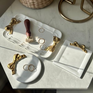 Puede incluir: Cuatro bandejas de cerámica blanca con detalles de lazo dorado. Las bandejas son perfectas para guardar joyas, llaves u otros objetos pequeños.