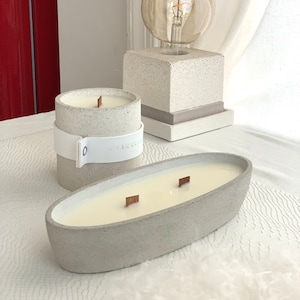 Puede incluir: Dos velas de hormigón gris con mechas de madera. La vela más grande es de forma ovalada y la más pequeña es cilíndrica. Ambas velas están sobre una superficie blanca.