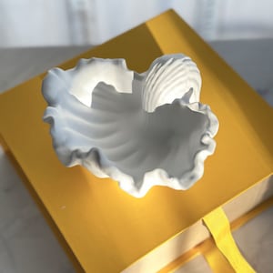 Puede incluir: Un plato de cerámica blanca con forma de concha marina con un borde rizado. El plato está sobre una caja amarilla con una cinta amarilla.