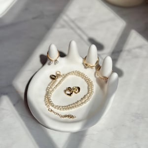 Op de afbeelding: Witte, handvormige sieradenschotel van keramiek met een parelketting, twee gouden ringen en een paar gouden hartvormige oorbellen.