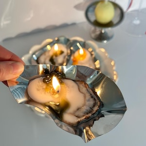 Op de afbeelding: Drie oester schelp kaarsen, twee zijn uit en één brandt met een kleine vlam. De kaarsen staan op een zilveren dienblad met een gegolfde rand.