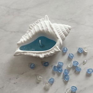 May include: A white seashell candle holder with a blue wax candle inside. The candle holder is surrounded by small blue and clear glass beads.