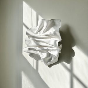 May include: White abstract wall sculpture with a flowing, draped design. The sculpture is made of a smooth, textured material and has a contemporary style.