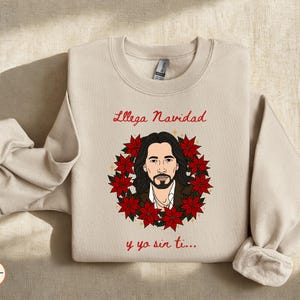 Op de afbeelding: Beige crewneck sweatshirt met een afbeelding van een man omringd door rode poinsettia's. De tekst "Llega Navidad y ya sin ti..." is in rood script boven en onder de afbeelding geschreven.