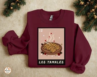 May include: Burgundy sweatshirt featuring a graphic of a plate of tamales with steam rising, surrounded by hearts and stars. The text "LOS TAMALES" is below the image. The number "12" is in the upper left corner of the graphic.