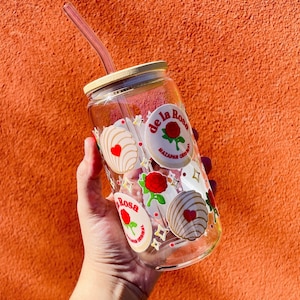 Op de afbeelding: Een heldere glazen fles met een houten deksel en een rietje. De fles is versierd met een patroon van rode rozen en witte en gouden snoepjes. De tekst "De la Rosa Mazapan Original" staat op de fles gedrukt.