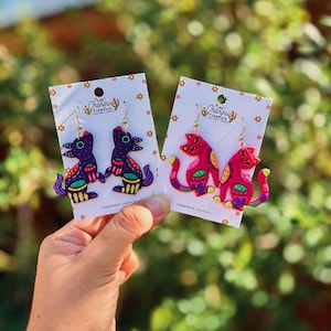 May include: Handmade earrings featuring colorful, stylized animal designs. One pair depicts purple and blue birds, the other, pink cats. Each earring is adorned with intricate patterns and the text "Las Changuis Tianguis Handmade Jewelry".