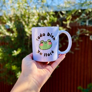 May include: A white ceramic mug with a light blue hue, featuring a cartoon frog design and the Spanish text "todo bien ya llor&eacute;." The frog has green skin and pink cheeks. The mug has a curved handle.