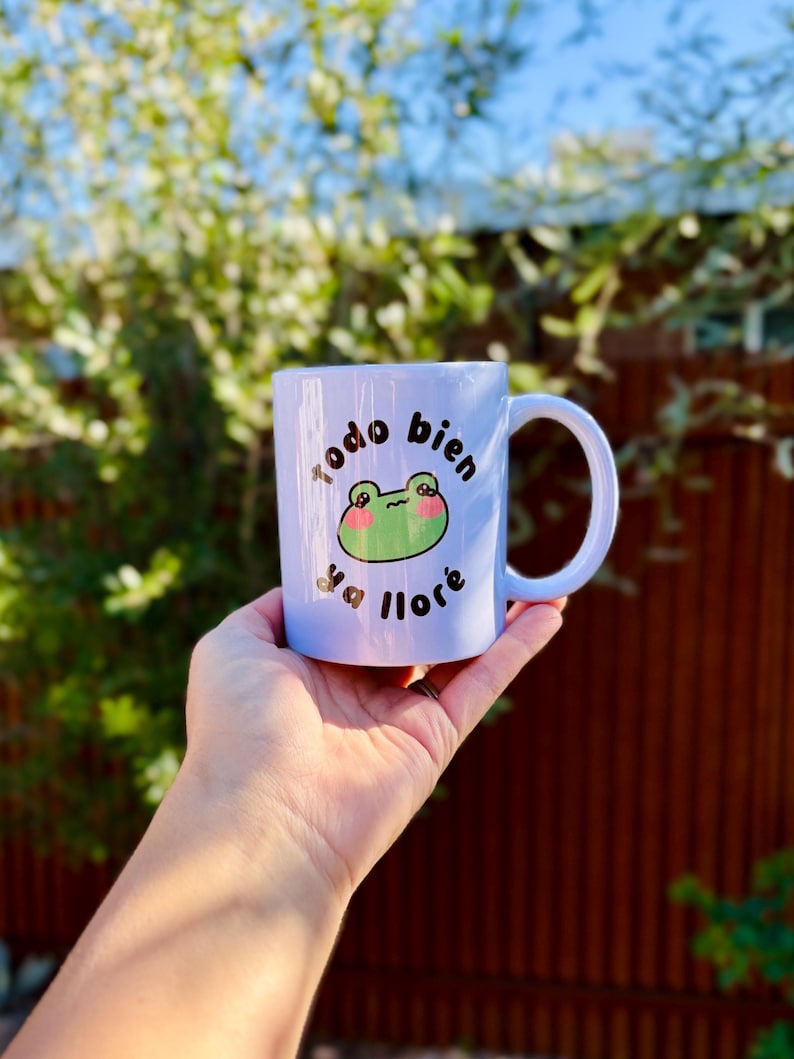 May include: A white ceramic mug with a light blue hue, featuring a cartoon frog design and the Spanish text "todo bien ya llor&eacute;." The frog has green skin and pink cheeks. The mug has a curved handle.