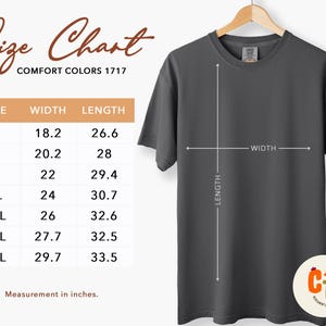 以下が含まれることがあります： 木製のハンガーに掛けられた濃い灰色のComfort Colors 1717 Tシャツ。画像には、Sから4XLまでのサイズのインチ単位の測定値を含むサイズチャートが含まれています。チャートはシャツの幅と長さを表示しています。