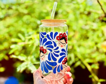 May include: A clear glass drinking cup with a bamboo lid and a glass straw. The cup is decorated with a blue and white design featuring a woman's face with a red flower crown. The cup is held in front of a blurred green background.