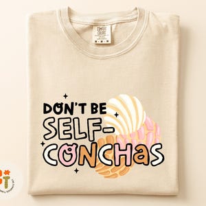May include: Beige Comfort Colors t-shirt with the phrase "Don't Be Self-Conchas" in black and pink lettering, with illustrations of conchas. The shirt has a crew neck and short sleeves. The shirt is folded.
