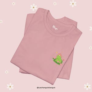 Może przedstawiać: R&oacute;żowy T-shirt z zieloną żabą w kapeluszu z grzyba, siedzącą na ł&oacute;żku z kwiat&oacute;w. Na koszulce znajduje się napis "Laschanguistianguis" u dołu.