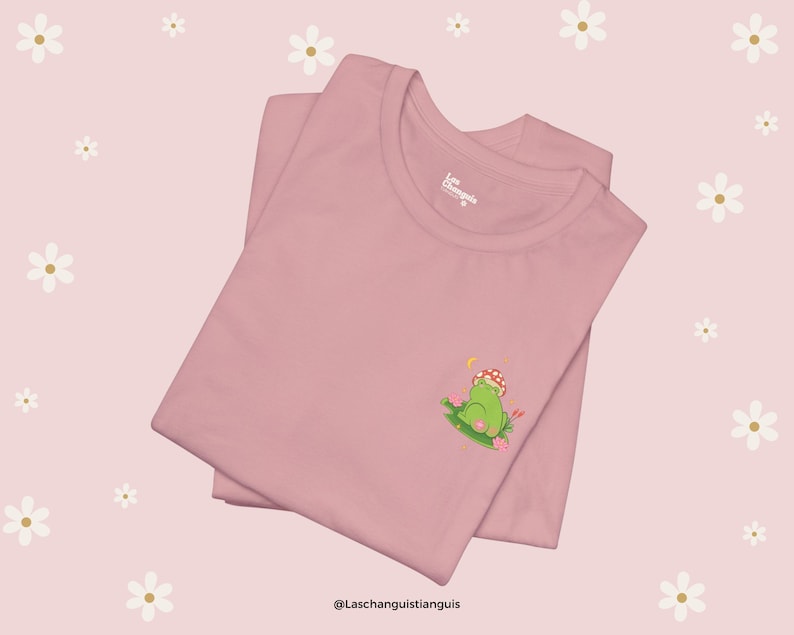 Może przedstawiać: R&oacute;żowy T-shirt z zieloną żabą w kapeluszu z grzyba, siedzącą na ł&oacute;żku z kwiat&oacute;w. Na koszulce znajduje się napis "Laschanguistianguis" u dołu.