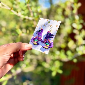 May include: A pair of handmade purple earrings with a unique bird-like design. The earrings feature gold-colored hooks and are adorned with colorful accents of red, green, and yellow. The earrings are displayed on a white card with the text "Las Changuis".