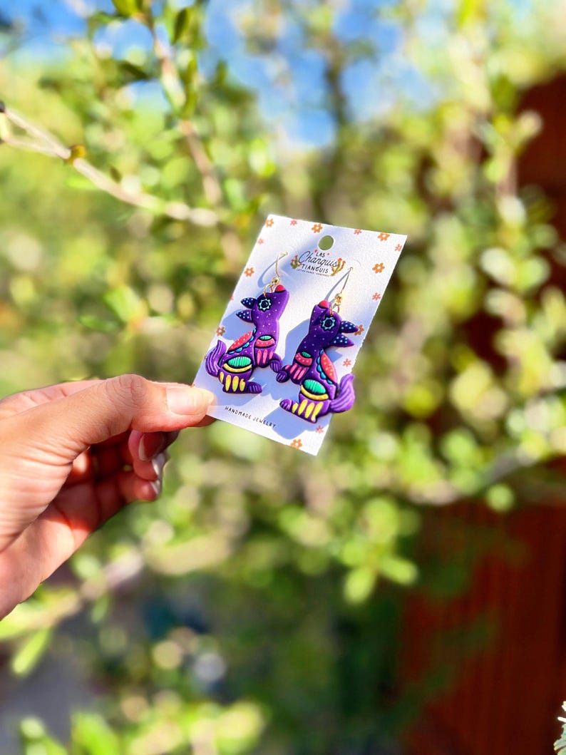 May include: A pair of handmade purple earrings with a unique bird-like design. The earrings feature gold-colored hooks and are adorned with colorful accents of red, green, and yellow. The earrings are displayed on a white card with the text "Las Changuis".