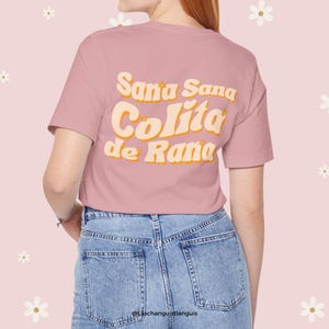 Może przedstawiać: R&oacute;żowy T-shirt z napisem "Sana Sana Colita de Rana" w retro czcionce. Tekst jest w kolorze ż&oacute;łtym i pomarańczowym.