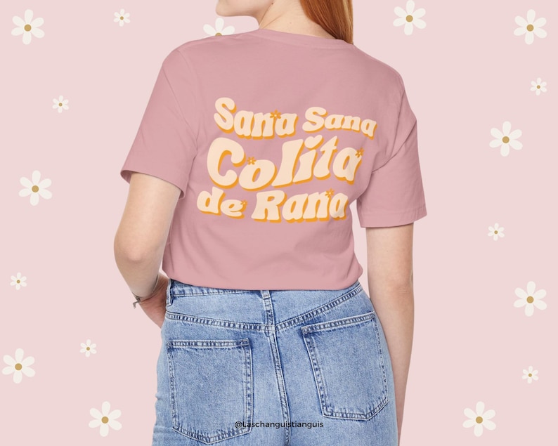 Może przedstawiać: R&oacute;żowy T-shirt z napisem "Sana Sana Colita de Rana" w retro czcionce. Tekst jest w kolorze ż&oacute;łtym i pomarańczowym.